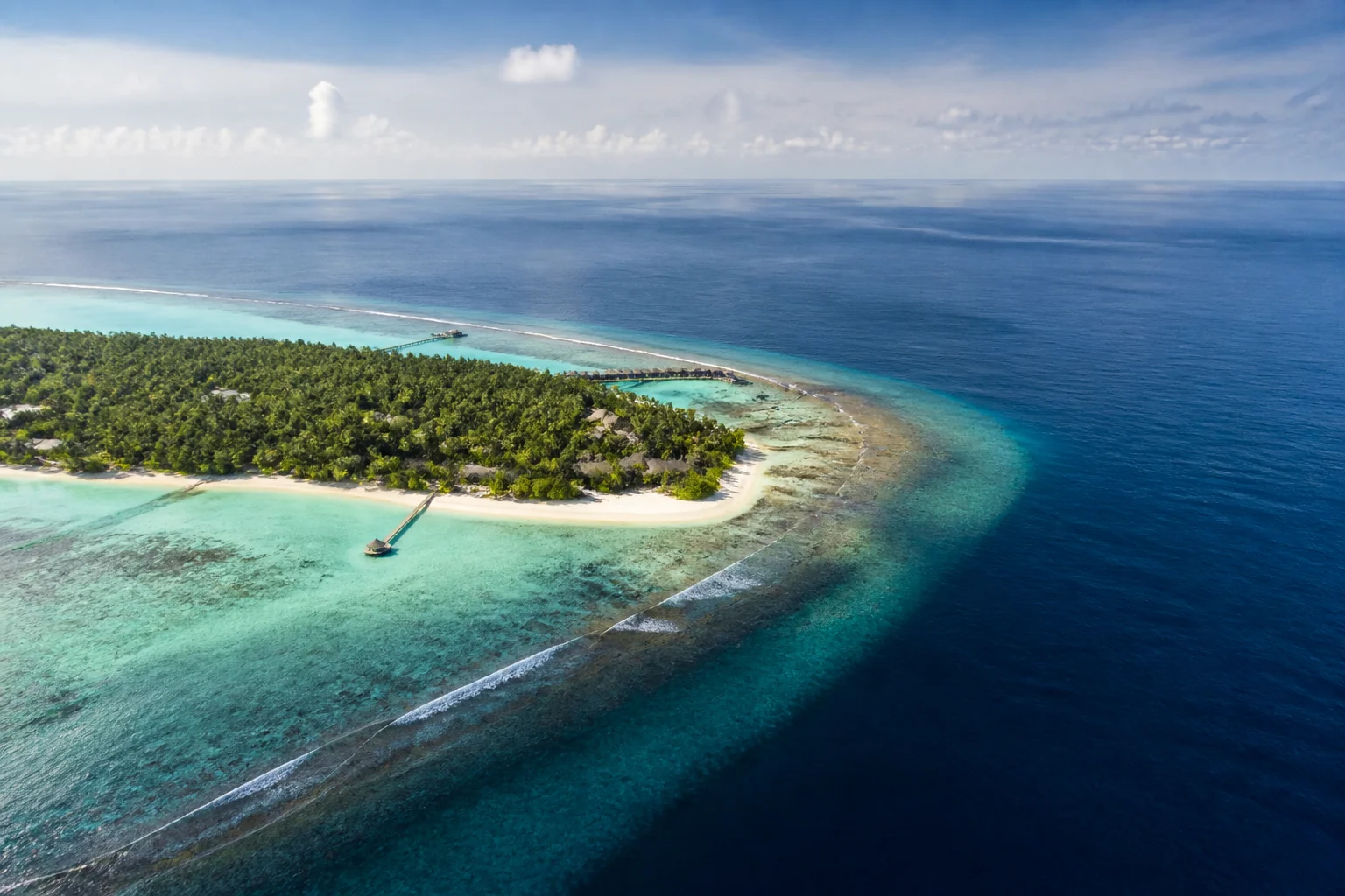 Maldivian resort island with pale lagoon, reef edge and deep outer water seen from above